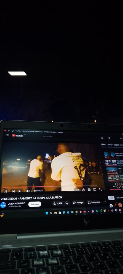A laptop displays a YouTube video titled "VEGEDREAM - RAMENEZ LA COUPE A LA MAISON." The video shows two men, one wearing a "ZIDANE 10" jersey, in what appears to be a night scene with other people. The YouTube interface is visible with options like "Partager" and "Enregistrer" and other suggested videos on the right side of the screen. The keyboard below, with a mix of English and Arabic characters, indicates the device is set to Arabic or supports both languages. A light source can be seen in the dark top-background.