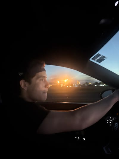 A dark image depicts a person's face in profile, illuminated against a backdrop of city lights. They appear to be in a car, looking out a window or sunroof. A raised arm is partially visible. The lighting suggests either dawn or dusk. The overall impression is one of contemplation or introspection amidst the urban landscape. The picture is upside down.
