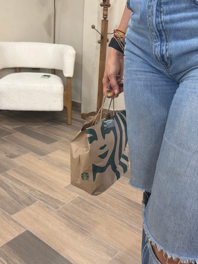 The image shows a person wearing light blue denim jeans, holding a brown paper Starbucks bag with green logos and handles. The person is wearing multiple bracelets on their left wrist. The background includes a light-colored tile floor in a herringbone pattern, a white upholstered bench or ottoman, and a wooden clothing rack. The location is likely indoors, but more context is needed to confirm the city of Villahermosa, Mexico.