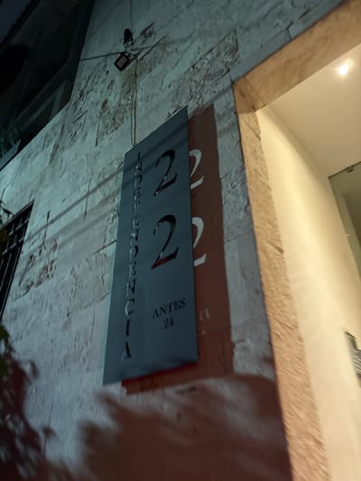 The image shows a sign for "Via Appia 22, Antes 24" attached to a stone wall. The location is likely Tizapan, Mexico. A light fixture is visible on the wall above the sign. The sign is in a dark color with white lettering. The wall appears to be textured and weathered. The angle of the photograph suggests it was taken from below the sign, looking upwards.