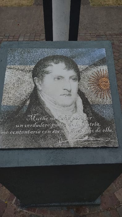 A memorial displays a grayscale portrait of a man with fair skin and dark hair, set against a faded Argentinian flag. Calligraphic Spanish text overlays the image, referencing patriotism and legacy. The memorial consists of a gray concrete pedestal with a plaque on top, situated on a brick-paved area. A black and white striped object is partially visible on the left, and dark shadows cast by the block enhance the memorial's somber tone.