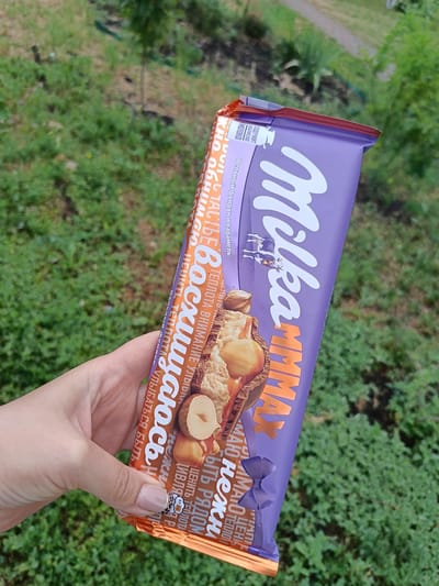 A person holds a purple and orange chocolate bar, identified as "Milka MMMAX," outdoors in Gubkin, Russia. The chocolate bar features text in Russian, with prominent orange lettering spelling out "Восхищаюсь" (I admire) and "MMMAX." The bar displays an image of chocolate, nuts, and caramel. The background consists of green grass and foliage. The person's hand and fingernails are partially visible, holding the chocolate bar.
