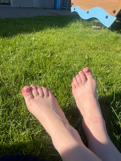 The image displays a close-up of two bare feet resting on bright green grass. The feet have a French manicure on the toenails, with a light pink polish and white tips. In the background, a portion of a playground structure with blue and wooden elements is visible, along with a building and a blue bin. The scene is lit by sunlight, casting shadows on the grass. The image appears to be taken outdoors, likely in a park or backyard. No specific location in Portlaoise, Ireland can be identified from the provided image.