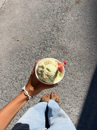 The image shows a person holding a cup of pistachio ice cream with a red plastic spoon. The ice cream is topped with cookie crumbs. The person is wearing light blue jeans and brown sandals. The person is standing on a concrete surface with a shadow to the right. The location is in Ciudad Santa Catarina, Mexico, which is a city located in the state of Nuevo Leon in the northeast of Mexico. The latitude and longitude coordinates (25.658, -100.438) place this scene in a residential neighborhood in the city of Ciudad Santa Catarina, Mexico.