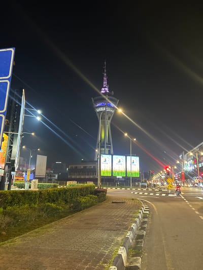 The image depicts a night scene in Alor Setar, Malaysia, focusing on the Alor Setar Tower (Menara Alor Setar). The tower, illuminated with purple and blue lights, stands tall in the background. In the foreground, a paved walkway runs alongside a road. The road is marked with white lines and has traffic lights, one of which is red. Several vehicles are visible on the road, including a motorcycle with a rider. Large digital billboards displaying advertisements are positioned near the tower. Streetlights cast long beams of light across the scene. The environment appears to be an urban setting with buildings visible further down the road. The walkway is bordered by neatly trimmed hedges.
