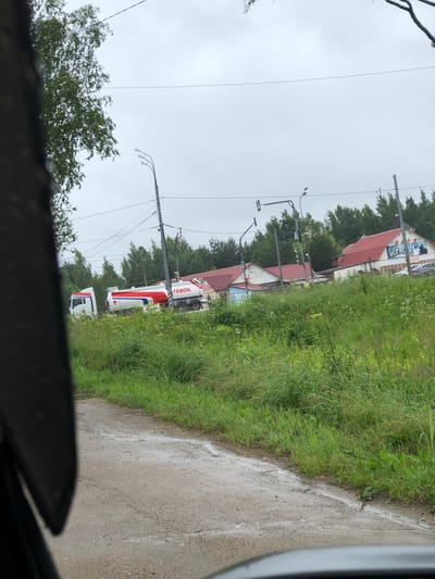 The image depicts a street scene in Safonovo, Russia, on a cloudy day. In the foreground, a portion of a dark vehicle's interior is visible, with the driver's side window framing the outdoor environment. The road surface appears wet, with puddles and a general dampness suggesting recent rain. Lush green grass and vegetation line the side of the road.
Further down the road, several buildings with red tiled roofs are visible. One building prominently features a large sign with text that includes "ТЕБОИЛ" and "БЕРЮЛ". A white tanker truck with red and white detailing is parked or moving on the road. Traffic lights and lampposts are present, indicating an urban or semi-urban setting. Trees and foliage are abundant in the background, forming a natural backdrop to the buildings and infrastructure. Several cars are parked near the buildings. The sky is overcast, contributing to the muted lighting of the scene.