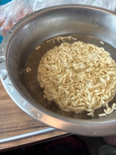 This image depicts a close-up, elevated view of an unidentifiable interior location in Tatarsk, Russia, focusing on a food preparation scene. A polished, silver-toned metallic bowl, possibly stainless steel, occupies the central and lower-right portions of the frame. Inside the bowl, a compacted block of light yellow, wavy instant ramen noodles is submerged in clear, hot water. Small bubbles are visible on the water's surface, indicating the noodles are in the process of cooking or soaking. Water droplets adhere to the exterior rim of the bowl on the left side. The bowl rests on a brown wooden surface, possibly a countertop or table, characterized by visible grain patterns. In the upper left background, a portion of a translucent plastic bag containing several orange-colored items, resembling carrots, is visible. Adjacent to the plastic bag, a light blue or teal surface is present. No individuals are visible in the scene.