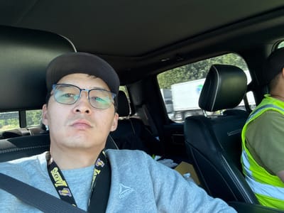 The image shows a man taking a selfie inside a vehicle. He is wearing glasses, a baseball cap, and a gray sweatshirt with a Reebok logo. A lanyard with a Los Angeles Lakers logo is around his neck. The car's interior is visible, including black leather seats and a sunroof. Outside the car, through the window, a highway with trees and a truck can be seen. Another person wearing a high-visibility vest is in the passenger seat. The location is identified as Ridgefield, United States of America.