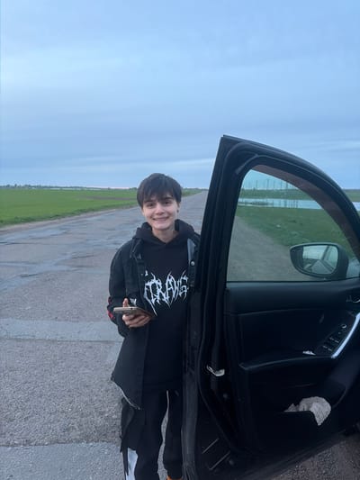 A person stands next to an open car door on a paved road in Azyakul', Russia. The person, dressed in a black hoodie with a graphic design and a dark jacket, is holding a smartphone. The landscape features a green field on one side of the road and a cloudy sky overhead. The side mirror of the vehicle reflects the surrounding environment.
