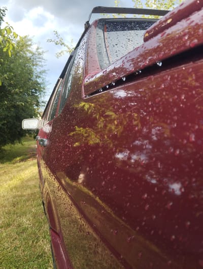 The image shows a close-up, low-angle view of the side of a dark red car covered in water droplets, parked outdoors. The car's paint reflects the surrounding environment, including green trees and a cloudy sky. A portion of a side mirror is visible on the left side of the frame, and a roof rack is present on top of the car. The ground beside the car is covered in green grass. The scene appears to be a rural or suburban setting, consistent with a location in Vinkivtsi, Ukraine, likely on a day with recent rainfall. There are no people or discernible actions taking place in the image.