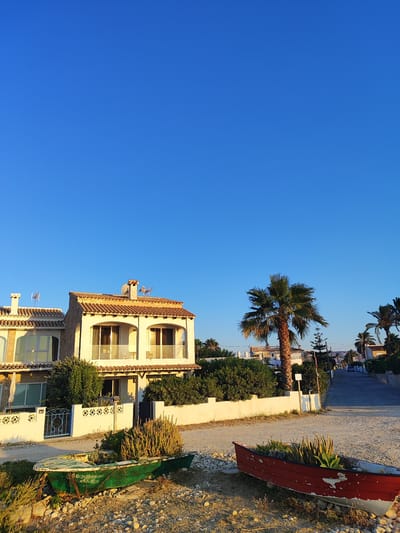 The image depicts a street scene in Urbanització Platja De L'almadrava, Spain, under a clear blue sky. In the foreground, two old, weathered boats are placed on a gravelly area, serving as planters for succulents and other greenery. The boat on the left is painted green, and the one on the right is red. Behind the boats, a white wall with decorative elements borders a property. A two-story house with a tiled roof, balconies, and arched windows is visible beyond the wall, with dense green foliage surrounding it. A tall palm tree stands prominently in the middle ground. Further down the street, other houses and palm trees are visible, and the road recedes into the distance, with a car parked on the side. The lighting suggests it is either early morning or late afternoon, casting a warm glow on the scene.