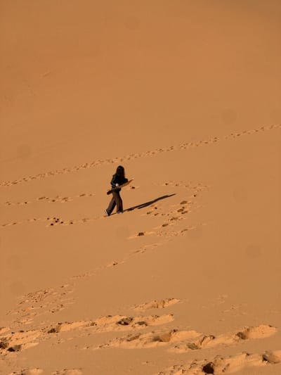 A high-angle, long shot depicts a single individual traversing a vast orange sand dune, likely within the vicinity of Rissani, Morocco, given the geographic context. The person, viewed from their left side, has dark, shoulder-length hair and is dressed in dark-colored attire, including a long-sleeved top or jacket and trousers. They wear light-colored footwear. The individual is in motion, walking from the lower left towards the upper right of the frame, ascending the dune. They carry a light-colored, rectangular board, approximately hip-width, horizontally in front of them with both hands. A distinct dark shadow extends behind and to the left of the person, indicating a strong light source from the upper right. The sand surface is covered with numerous human footprints, suggesting prior passage. The dune's texture appears soft and undulating. The sky is not visible; the frame is dominated by the uniform orange hue of the sand.