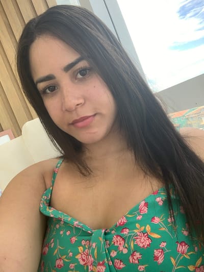 A female subject is depicted in a head-and-shoulder portrait, positioned indoors in Lauro de Freitas, Brazil. She has long, dark hair, dark eyes, and visible freckles across her nose and cheeks. Her lips are a natural tone. The subject is wearing a green top with pink and red floral patterns, featuring thin shoulder straps and a knotted detail at the bust. Her right shoulder and upper arm are partially visible on the left side of the frame. The background to the left consists of light-toned vertical wooden paneling, a portion of a cream-colored furniture arm, and a partial view of a light-colored picture frame. To the right, a large window shows a bright, cloudy sky and, in the mid-distance, a building with a terracotta-tiled roof and light-colored walls. Natural light illuminates the scene, appearing to originate from the right. The subject maintains direct eye contact with the camera; no other individuals or specific actions are present.