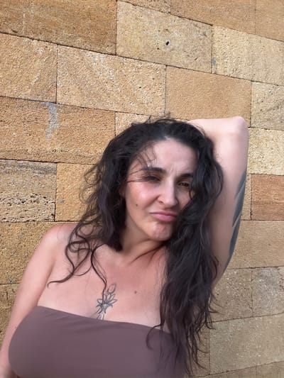 The image shows a woman lying on a textured, tan-colored tiled surface. She has long, dark, wavy hair that falls around her face and shoulders. Her left arm is bent, with her hand behind her head. She is wearing a brown strapless garment. A tattoo is visible on her chest, featuring a stylized floral or celestial design. Another tattoo is visible on her left arm. Her facial expression is somewhat ambiguous, with her eyes partially closed and her lips slightly parted, suggesting a relaxed or pensive mood. The lighting suggests it is daytime, with shadows indicating sunlight. The tiled surface appears to be outdoors, possibly a patio or walkway. There is no visible text in the image.