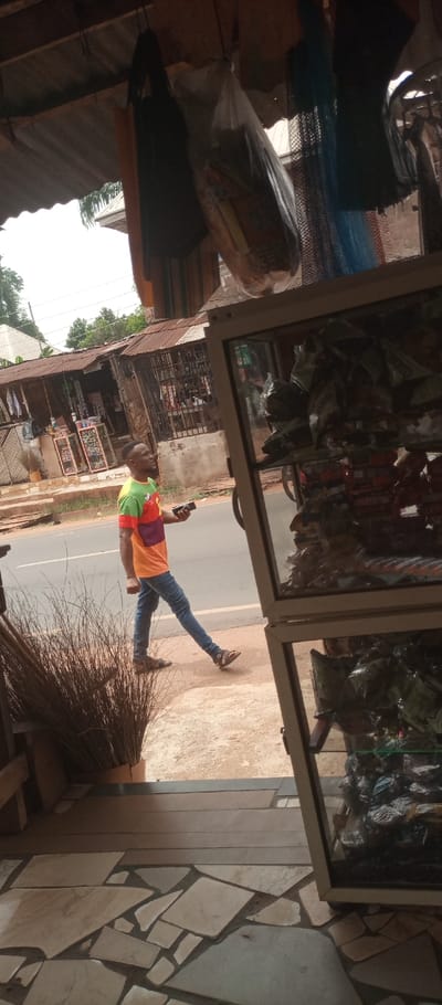 The scene is captured from inside a roadside shop or stall in Umuoji, Nigeria, looking out onto a street. The foreground displays an irregularly tiled floor in light tones. To the left, a vertical bundle of dried, dark brown reeds or brooms stands in a container. To the right, a two-tiered, light-framed glass display cabinet filled with numerous small, dark-packaged items occupies the frame. Overhead, various items hang from a corrugated metal ceiling, including a dark bag, an orange textile, a white plastic bag containing provisions, and a blue mesh item.
In the midground, an adult male walks from left to right on a reddish-brown dirt verge adjacent to a grey asphalt road. He is dark-skinned, with short dark hair and a beard. He wears a multi-colored t-shirt featuring green, red, orange, purple, and a yellow patch, blue jeans, and brown open-toed sandals. He holds a dark-colored smartphone in his right hand, extended forward, and his gaze is directed forward and slightly to his right.
Across the road, multiple single-story commercial buildings with corrugated metal roofs are visible. These include a structure with a white, angular roof component on the left and another with a dark metal gridded storefront displaying various goods. Green foliage and trees are present in the distant background under a bright sky.