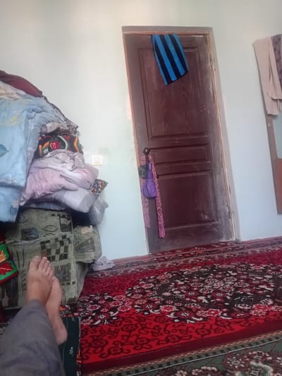 The image depicts an indoor scene within a room, likely in Galaosiyo, Uzbekistan. In the foreground, a person's feet, clad in grey pants, are visible resting on a carpeted floor. The carpet features a prominent red base with intricate floral patterns in white and black. To the left, a large, multi-layered stack of blankets and pillows is piled on top of what appears to be a sofa or seating unit. The seating unit itself is covered in a patterned fabric, primarily green with darker checkered sections. A light switch is visible on the wall behind the piled blankets. The background is dominated by a dark wooden door, slightly ajar, with a blue and black striped cloth hanging from its top edge and a purple umbrella and pink scarf dangling from the doorknob. To the right of the door, a section of a wall is visible, with two pieces of draped clothing hanging from what might be a rail or hook. The overall impression is of a lived-in, possibly untidy domestic space.