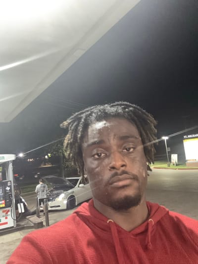 A man in a red hoodie is taking a selfie at a gas station in Arlington, Texas, USA. The gas station has a 7-Eleven store, and a sign for St. Michael is visible in the background. A car is pulled up to the pump, and a person appears to be working on the engine. It is nighttime.