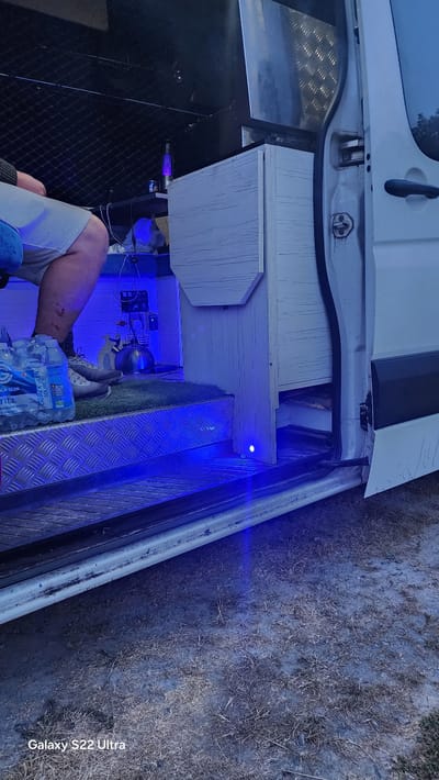 The image shows the interior of a van, specifically the driver's side entry with the door open, parked on a grassy and dry ground surface, possibly a campsite or rural area. The van's interior is partially fitted out as a living space, with a white wooden cabinet visible on the right. Blue LED lights are illuminating the floor area of the van and the immediate exterior. A person's legs and lower torso are visible on the left, wearing light-colored shorts and athletic shoes. They are seated within the van. In front of the person, a six-pack of "CARRICK GLEN" mineral water bottles is placed on a silver, diamond-plated surface. Further inside the van, a small kettle is visible on a shelf, along with some electrical equipment and what appears to be a bottle of cleaning spray. The lighting suggests it is either dusk or night. The lower-left corner of the image displays the text "Galaxy S22 Ultra", indicating the device used to capture the photograph. The location within the city of Cemaes, United Kingdom, is not discernible from the visual information present in the image.