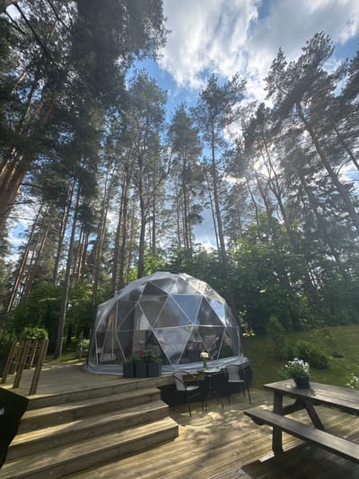 The image depicts a geodesic dome glamping tent situated in a wooded area. The dome is made of transparent panels and has a wooden deck extending from it. There are several chairs and a table on the deck, as well as a picnic table with benches. The surrounding environment is a forest with tall pine trees. The scene appears to be set in Priedesmuiža, Latvia, suggesting a tranquil, nature-focused accommodation. No people are visible in the image. The lighting indicates it is daytime with sunbeams filtering through the trees.