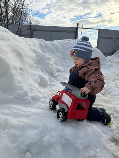 This image captures a young child kneeling in a snowy outdoor environment within the city of Уром, Russia. The child, clad in a grey and white striped knit beanie with a pompom and a "Cars" character patch, a brown winter jacket with dark tree or paw print patterns, and dark blue or black snow pants over lime green accented shoes, is observed looking downwards and to their right. The child's left hand rests on a large red plastic toy fire truck, which has black wheels and a grey ladder apparatus on its top. The ground is covered extensively with granular white snow, forming a significant accumulation or snowbank that rises behind the child and extends to the left. In the background, a light-colored corrugated metal fence or wall is visible, with a few bare tree branches extending from the upper left and a wooden post positioned to the right of the fence. The sky above is partially clear, displaying patches of blue interspersed with white clouds.