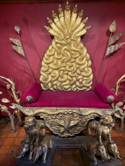 The image displays a throne situated in Lalitpur, Nepal. The throne is decorated with an elaborate golden backrest that appears to be styled with multiple snake heads and a pattern of interwoven serpentine forms. The seat of the throne is upholstered in a magenta-colored material with cylindrical cushions on either side. The throne also features golden animal figurines at its corners. Spears flank the backrest, and the throne is set against a red wall, with a tiled floor visible at the bottom of the image.