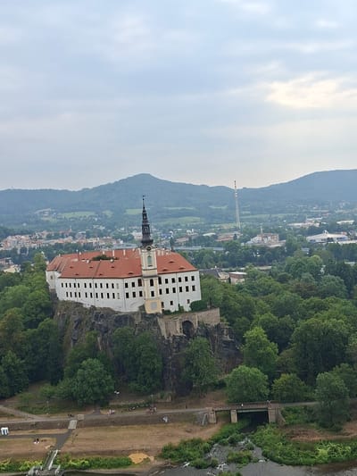 The image captures an elevated view of Děčín Castle, a prominent landmark in Děčín, Czechia. The castle, with its white facade and red-tiled roof, is situated atop a rocky outcrop, surrounded by lush green trees. A tall, dark spire with a clock face rises from the castle's center. In the background, a sprawling cityscape is visible, characterized by a mix of residential buildings, industrial structures, and rolling green hills. A tall, striped chimney stack stands out against the sky in the middle ground. A river flows in the foreground, with a bridge crossing it and a pathway running alongside it. Several people are scattered throughout the scene, some walking or cycling along the path, and others appearing to be relaxing on the grassy embankment. The sky is overcast with diffused light.