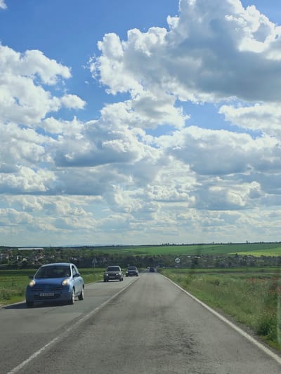 The image depicts a rural road in Bulgaria, seemingly near the city of Kumanovo, as the license plate "B 6238 BC" is visible on the lead blue car, indicating registration in that area. Several vehicles are moving along the road, with the blue car in the foreground. The road is bordered by green fields on one side and a line of trees and a village on the other. The sky is predominantly cloudy, with patches of blue visible.
