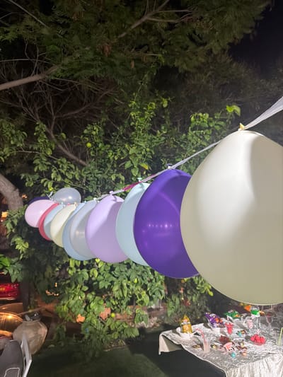 The image displays a festive outdoor scene at night, likely in Kochav Yair Tzur Yigal, Israel. The central focus is a vertical string of helium balloons, ascending towards the top of the frame. These balloons feature a pastel color scheme, including off-white, purple, light blue, lavender, light grey, cream, and light pink, arranged in a descending size order from the top.

The balloons are set against a backdrop of lush, dark green trees and bushes, illuminated by artificial lighting that highlights the foliage and the balloons while the sky remains dark. Small, round green fruits are visible on some of the branches, suggesting the presence of citrus trees. In the upper right corner, a silvery, crinkled fabric is draped, from which numerous small, colorful ornaments or decorations, possibly party favors or toys, are suspended. The overall impression is a celebratory event or party held outdoors during the evening. No visible text is present in the image.