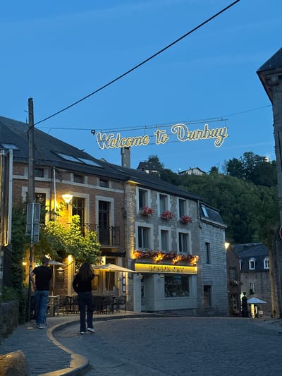An outdoor, medium shot captures a cobblestone street in Durbuy, Belgium, illuminated by the soft glow of streetlights and a neon sign that reads "Welcome to Durbuy." The scene is set during twilight, with a clear, dark blue sky. Buildings of brick and stone line the street, some with windows and balconies adorned with potted flowers. A couple walks away from the camera down the curved street. One person, a male, wears a black t-shirt and blue jeans. The other, a female, wears a black top and dark pants. They are walking past a building with outdoor seating and umbrellas, suggesting a restaurant or cafe. The neon sign, suspended above the street, is the focal point, casting a warm yellow light. The architecture is old-world European, with a steep hill covered in lush green trees rising behind the buildings. A streetlight on the left side of the image casts a warm yellow glow on the surrounding foliage.