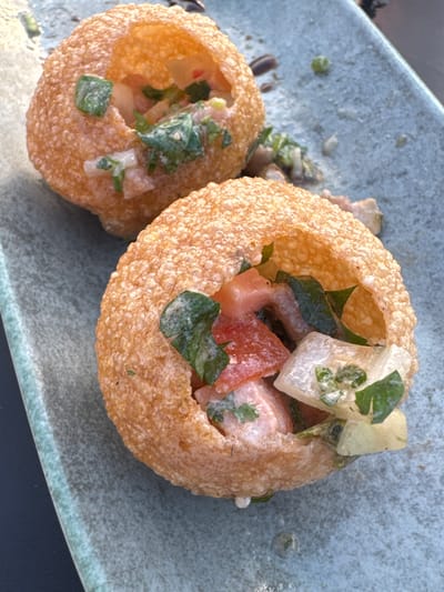 The image displays two golden-brown, spherical, fried pastries, consistent with *pani puri*, presented on a light blue-grey rectangular ceramic plate with a speckled texture. The scene is located in Ness Ziona, Israel. Each pastry is open at the top, revealing an internal filling. This filling consists of diced red components (likely tomatoes), irregularly shaped white or pale-colored pieces (possibly potatoes or onions), and distinct green leafy fragments (consistent with herbs like cilantro or mint). A light-colored, possibly creamy or watery, dressing coats the interior ingredients. The pastry in the upper-left is partially visible, while the one in the lower-right is more centrally positioned. The plate surface shows minor debris, including small green specks and slight liquid traces. A dark, unidentifiable surface serves as a background element on the left and lower-left portions of the frame. No individuals are present in the image.