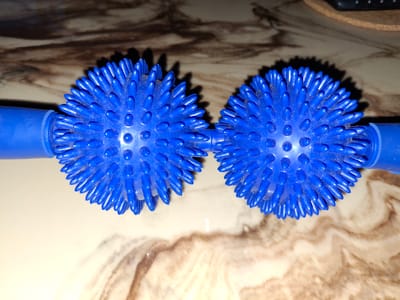 Two bright blue, spiky massage balls are positioned side-by-side on a marbled surface, connected by a blue rod. The balls are spherical and covered in numerous short, pointed protrusions, resembling sea urchins. Each ball has a diameter of approximately 5 centimeters. The marbled surface exhibits swirling patterns of brown and beige, with some areas appearing lighter and others darker, creating a natural stone-like texture. The lighting casts distinct shadows behind the massage balls, indicating a strong light source from the top-left. The overall scene is a close-up, top-down view of the massage balls. There is no indication of the city of Åsgårdstrand, Norway, or any specific location within it.