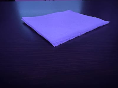 A single, folded paper napkin, colored light purple, rests on a dark, wooden surface. The napkin is positioned diagonally in the frame, with its top-left corner closest to the viewer and extending towards the bottom-right. The lighting is dim and casts a blueish-purple hue across the scene, suggesting an artificial light source. The texture of the napkin is visible, appearing somewhat rough and creased, indicating it has been handled. The wooden surface beneath it has a subtle wood grain pattern that is mostly obscured by the dark, reflective lighting. There are no people, animals, or other objects present in the image. The image does not contain any information that would allow for a specific location in Mastaga, Azerbaijan to be identified.