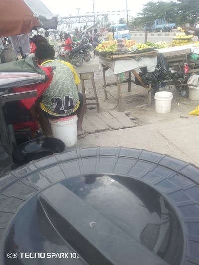 This image depicts an outdoor market scene in Adesanya, Nigeria. The foreground is dominated by the lid of a large, dark plastic container, possibly a water tank, reflecting the sky and surrounding environment.

In the mid-ground, a person wearing a green and yellow patterned shirt with the number "24" is seated on a white bucket. They are facing away from the camera, and their legs are visible, wearing sandals. Behind them, part of a stall with a red cloth and other items is seen.

Further back, several other individuals are present, some appearing to be vendors or shoppers. A person in a red shirt is seen on a motorcycle, and others are visible around stalls laden with fruits, including what appear to be apples, grapes, and lemons. A blue truck and other vehicles are in the background, suggesting a busy roadside market.

The setting appears to be a market located alongside a road, with various stalls and temporary structures. The ground is dusty and uneven, with some wooden planks laid out. The weather appears to be overcast or hazy, and the time of day is likely daytime. The overall impression is that of a lively, everyday market in Nigeria.

The visible text in the image is "TECNO SPARK 10c" located at the bottom left corner.