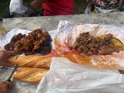 A group of people are gathered around a concrete table, enjoying a meal outdoors. The table is laden with food, including several baguettes, skewers of grilled meat, and chopped steak served on parchment paper. Two people are visible in the background, one wearing a red shirt and the other a floral-patterned top. A hand is shown slicing into a baguette with a knife. The environment appears to be a park or recreational area, with greenery visible in the background. The scene suggests a casual outdoor dining experience.