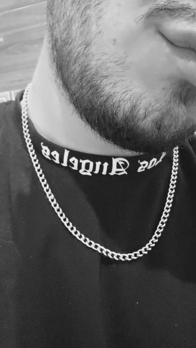 A close-up, black and white photograph depicts a portion of a person's face and torso. The person has a beard. A silver chain is visible around the person's neck. White lettering, written in a stylized font, is visible on the person's black shirt. The text reads "St. Jude & Co.". No location or city information is discernible.