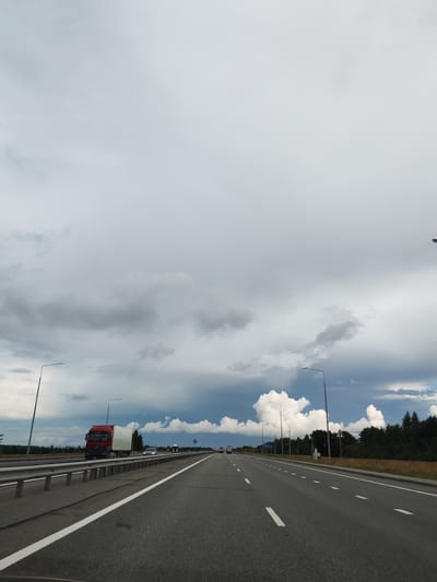 The image depicts a perspective from a moving vehicle on a highway, likely in or near Vasiukovo, Russia. The road is asphalt with clear white lane markings. A red and white semi-trailer truck is visible in the left lane, traveling in the same direction as the viewer. Several other vehicles are seen further down the highway. Tall streetlights line the side of the road. The sky is heavily overcast with dramatic dark clouds, suggesting an impending storm or recent rainfall. The landscape on either side of the highway consists of trees and fields.