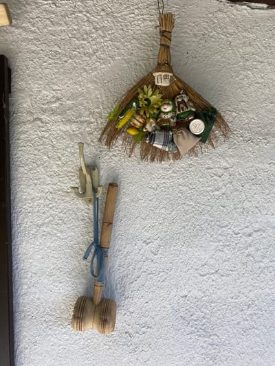 The image shows a rustic display on a textured white wall, likely outdoors given the natural light. The main subjects are a decorative broom and a wooden mallet hanging on the wall. The broom is adorned with various small figurines, including what appear to be traditional Japanese Daruma dolls and small bags, along with miniature produce like corn and gourds, suggesting a festive or harvest theme. A small, white, house-shaped ornament with Japanese characters (likely "田上" or "Tagami") is also attached to the broom.
To the right of the broom, a wooden mallet with a blue ribbon tied around its handle is mounted on a light-colored, vintage-style hook. The mallet has a distinct, grooved head.
The overall setting appears to be rural, consistent with the provided location context of a rural council in Belarus. The lighting suggests daytime, and the textured wall adds a sense of age and character to the scene. There are no people visible, and the objects are stationary, indicating a decorative arrangement rather than an active event. The white circular object on the broom appears to have some inscribed text and a symbol, but it is too small and blurry to be definitively read.