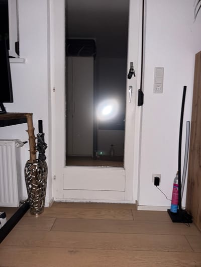The image depicts an indoor scene, likely a hallway or entryway, in a residential setting. The dominant feature is a glass-paned door, partially open, leading into a dimly lit interior space. To the left of the door, a white radiator is visible, and next to it, a decorative vase holds a natural branch and a black umbrella. A thin, dark metal bar runs along the floor in front of the vase and extends out of frame to the left.

The wall to the right of the door is white and features a light switch and an electrical outlet with a black adapter plugged in, to which a thin black wire is connected. A tall, black object, possibly a slim lamp or decorative item, stands vertically to the right of the outlet. Further to the right, a pink and blue aerosol can is positioned on a black base, with wires trailing from it. A section of a wooden surface is visible at the far right edge of the image. The floor is composed of light-colored wooden planks.

The scene appears to be captured in low light conditions, with a bright, circular reflection visible on the glass door, suggesting a light source from within the interior. There is no indication of the city of Langen bei Bregenz, Austria, in the visual elements of the image.