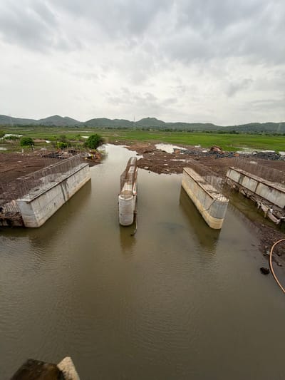 The image depicts a construction site for a bridge in Saphale, India. The scene shows several concrete pillars and beams emerging from murky water, indicating that the bridge is under construction over a river or canal. The surrounding landscape is a mix of undeveloped land with piles of construction materials and lush green fields with rolling hills in the background. The sky is overcast with heavy clouds, suggesting a possibly rainy or humid day. There are no visible people actively working in the immediate foreground of the construction, but a single figure appears to be present near one of the pillars. The overall impression is of a large infrastructure project in progress within a natural, undeveloped setting.