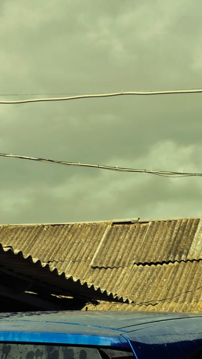 The image displays a close-up view of corrugated roofing material in an outdoor setting, under a muted, overcast sky. Two power lines are visible in the upper portion of the frame, running horizontally across the sky. Below the sky, the aged, weathered corrugated roof occupies the majority of the image, showing signs of wear and tear. In the foreground, a portion of a blue vehicle is visible, with its shiny surface contrasting with the rough texture of the roof. The scene appears to be in Troițcoe, Moldova, suggesting a utilitarian or residential exterior. There are no people or explicit actions depicted, the image focuses on the static elements of the environment.