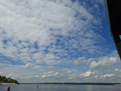 The image depicts a wide, calm body of water, likely a river or lake, under a bright blue sky dotted with numerous white, fluffy clouds. On the left bank, a verdant hill slopes down to the water, topped with several buildings, including what appears to be a large house or mansion surrounded by trees. A red buoy is visible in the foreground on the water, and another smaller, red and white buoy is further in the distance. Across the water, the opposite bank is lined with a distant cityscape and a dense line of green trees. In the upper right portion of the frame, a dark, angled structure, possibly part of a boat or bridge, partially obstructs the view. The overall scene suggests a sunny day with mild weather, likely in the city of Verkhny Uslon, Russia, given the context of the prompt.