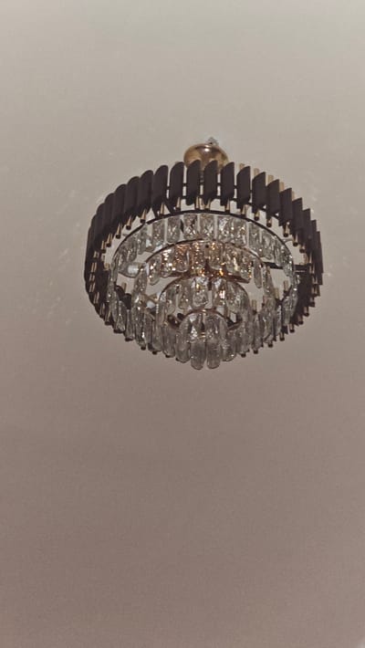 A multi-tiered chandelier hangs from a white ceiling. The chandelier features a dark brown outer rim with a scalloped edge, and several layers of clear, faceted crystal droplets. The upper portion of the chandelier has a golden fixture. The overall lighting is dim, with faint shadows cast on the ceiling. The image does not contain any information about the city of Gurlan, Uzbekistan.