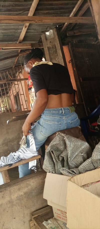 An individual, viewed from the rear, is seated within a market stall or shop in Umuoji, Nigeria. The person wears a black short-sleeved top exposing the lower back and light blue denim jeans with a green belt; dark braided hair is pulled up. In the left hand, a white fabric with black linear patterns is held. The individual is seated on a dark brown cushioned stool. To the right of the seated person, a crumpled black and white checkered fabric rests on a surface next to stacked cardboard boxes. One box displays partial red text, including "IKEJA, LA" and "STORE IN A C." The immediate environment features a wooden plank or counter. In the background, a metal-barred opening, a light-toned wall, and shelves displaying merchandise are visible on the left. A partial blue plastic chair is visible on the right background. The overhead structure consists of a corrugated metal roof supported by wooden planks and beams, indicating an informal commercial establishment.