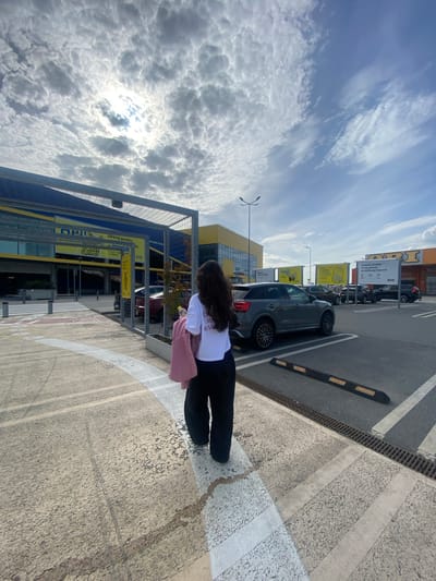 A person wearing a white t-shirt and black pants walks away from the camera in an outdoor parking lot. They are carrying a pink bag over their shoulder and a pink jacket draped over their arm. The person is walking on a textured concrete surface with white painted lines. In the background, a large yellow and blue building, identified as an IKEA store in Bielany Wrocławskie, Poland, is visible. The building has a large canopy structure. Several cars are parked in the lot, and a billboard with text in Polish is also present. The sky is partly cloudy, with the sun visible through a break in the clouds.