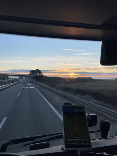 A sunset viewed through a vehicle's front window reveals contrails streaking across a blue sky. The highway stretches forward, marked with "260m" and white lane lines. A navigation device displays a route with "100 m" and "82" visible, leading towards "La Almolda," as noted at 16:33, 32:01, and 2836. The reflection of the sunset is also on the front window of the vehicle.
