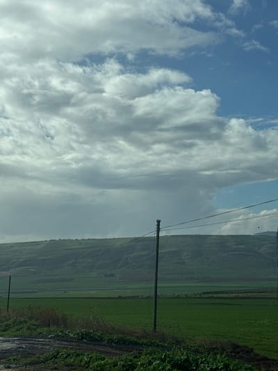 The image displays a panoramic view of a rural landscape near Tiberias, Israel, under a partly cloudy sky. The upper two-thirds of the frame are filled with a dynamic arrangement of cumulus clouds, ranging in color from dark grey and slate to bright white, interspersed with patches of clear blue sky visible predominantly in the upper right quadrant. A large, dark grey cloud mass is prominent in the mid-left.
A continuous, verdant hill range forms the horizon line across the mid-ground, characterized by a gently undulating crest and slopes covered in varying shades of green vegetation, consistent with cultivated fields or natural grasslands. Below this elevated terrain, expansive, flat agricultural fields, predominantly a vibrant green, stretch towards the foreground. In the mid-right foreground, a single, dark-colored utility pole stands vertically, with multiple power lines extending horizontally from its upper section. The immediate lower-left foreground shows a dark, muddy dirt path or ground surface, with sporadic patches of darker green, low-lying vegetation. The scene is devoid of human figures, vehicles, or active wildlife.