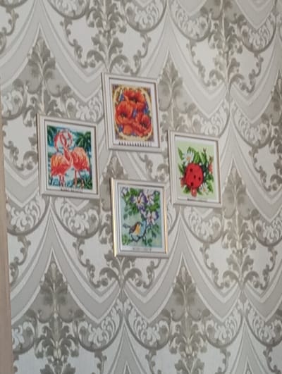 An interior wall in a residence located in the city of Яромаска, Russia, is depicted. The wall is covered with light-toned wallpaper featuring a repeating, ornate damask-style pattern in a slightly darker grey hue. Four square, light-colored framed artworks are affixed to the wall in a staggered, asymmetrical cluster. The artwork appears to be cross-stitch or diamond painting. The top-most frame displays red poppies with green leaves and blue accents. To the left and slightly below is a frame containing an image of two pink flamingos in a tropical setting. To the right of the poppy frame is a piece depicting a red ladybug on green foliage with small white flowers. Centered below the flamingo and ladybug frames is a fourth piece portraying a small brown and orange bird perched on a branch with purple flowers. No individuals, actions, or interactions are visible within the scene.