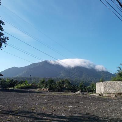 The image shows a view of a mountainous landscape under a clear blue sky. In the foreground, there is a dark, gravelly area with some scattered debris and vegetation. A low stone wall is visible on the right side of the frame. In the midground, there are lush green trees and foliage. Dominating the background is a large mountain, partially shrouded by a band of white clouds that stretches horizontally across its summit. Several power lines are strung across the sky, running diagonally from the upper left to the upper right of the image. The scene appears to be a rural or semi-rural area, possibly in Dolat Rayat, Indonesia, given the lush vegetation and mountainous terrain. No people are visible in the image.