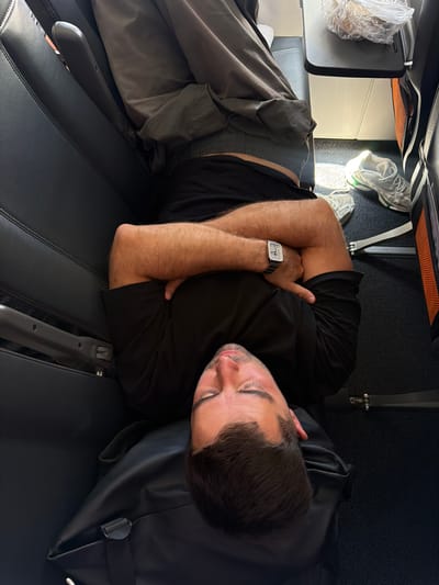A man is sleeping on an airplane with his arms crossed over his chest. He is wearing a black t-shirt and black shorts. A gray jacket is draped over his legs, and a black bag is placed under his head, serving as a pillow. White sneakers are visible on the floor next to the man's feet. A small tray table with a plastic-wrapped item is also present. The scene suggests a travel context.