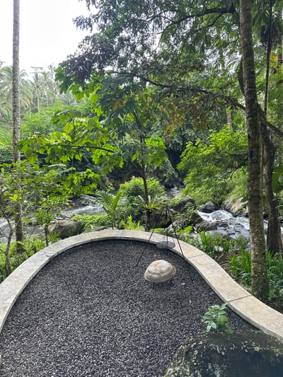 A medium-shot, landscape-oriented image depicts a portion of an outdoor seating area located near a waterfall in Licin, Indonesia.

The foreground features a curved concrete wall enclosing an area filled with small, dark gravel. A light-colored basin sits on the gravel surface, near a metal chair. The area is surrounded by dense vegetation, including trees and ferns. A waterfall cascades down rocks in the background. The scene is captured during daylight hours.